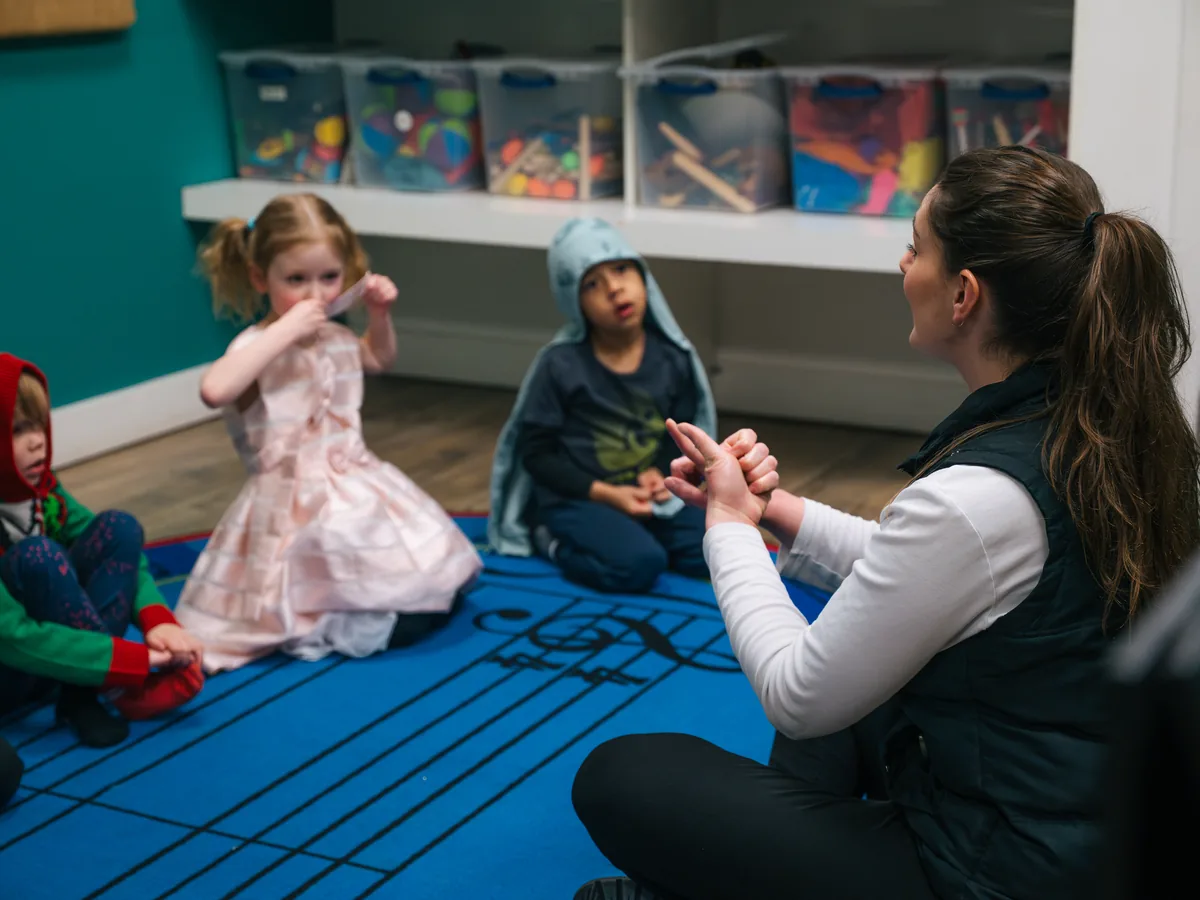 Teacher seated on a music staff mat showing a hand sign to small children during a Tunes and Tots class at Resonate